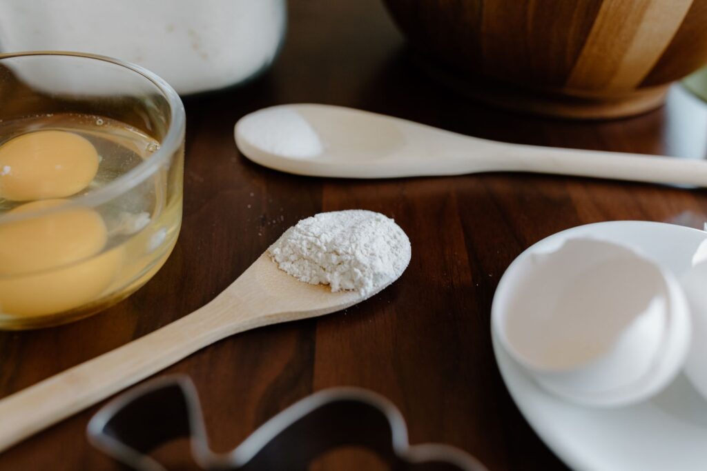 Spoonful of white baking powder on a wooden spoon beside cracked eggs and a bowl, ready for gluten-free baking