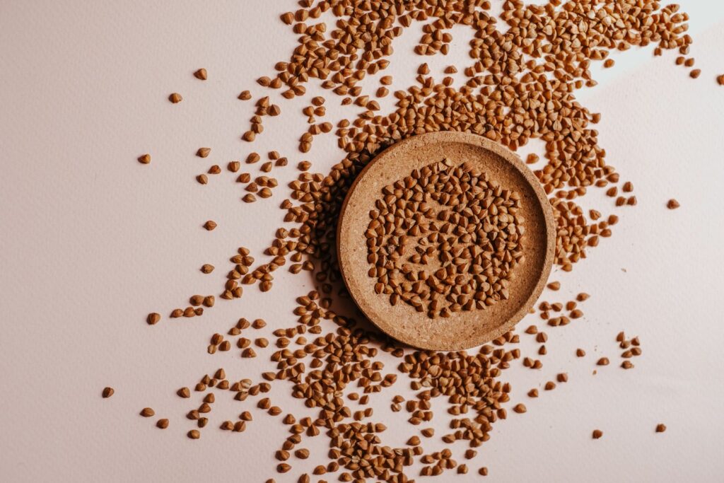 Buckwheat groats in a small wooden dish with more groats scattered across a light surface, illustrating the gluten-free seed