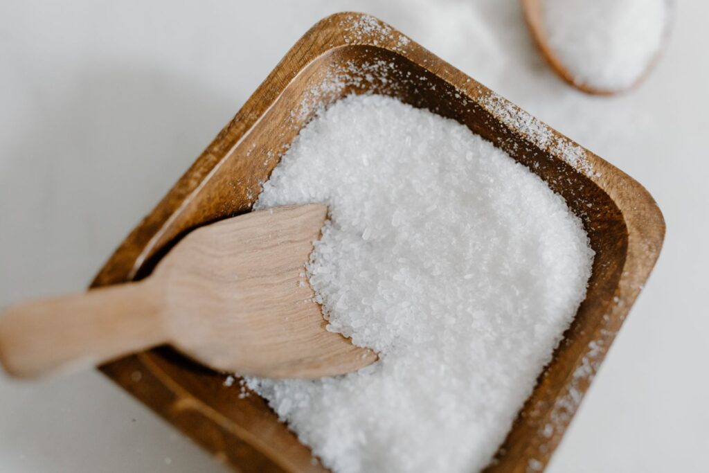 Fine white crystalline powder in a wooden dish with a small wooden scoop, illustrating monosodium glutamate (MSG).