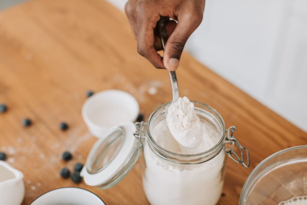 Hand scooping fine white flour with a spoon from a glass jar, a gluten-free baking ingredient scene with blueberries nearby