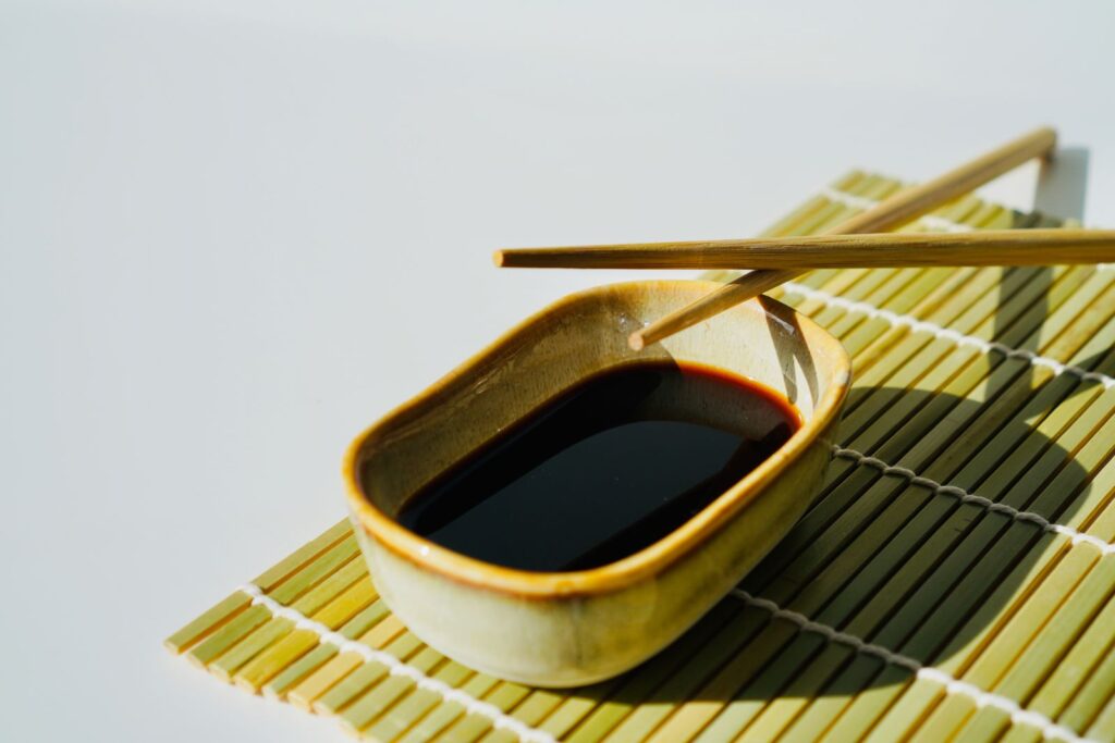 Small ceramic dish of dark soy sauce on a bamboo mat with chopsticks resting across it, ready for a gluten-free dipping check