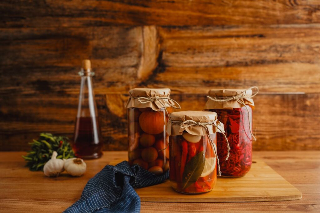 Jars of vegetables pickled in vinegar beside a bottle of vinegar and fresh garlic on a rustic wooden table