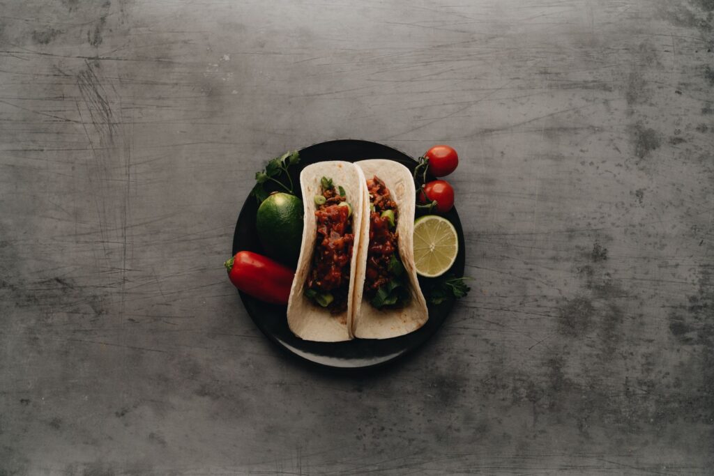 Two soft flour tortillas filled with seasoned meat and salsa on a dark plate with lime, tomatoes and chili pepper