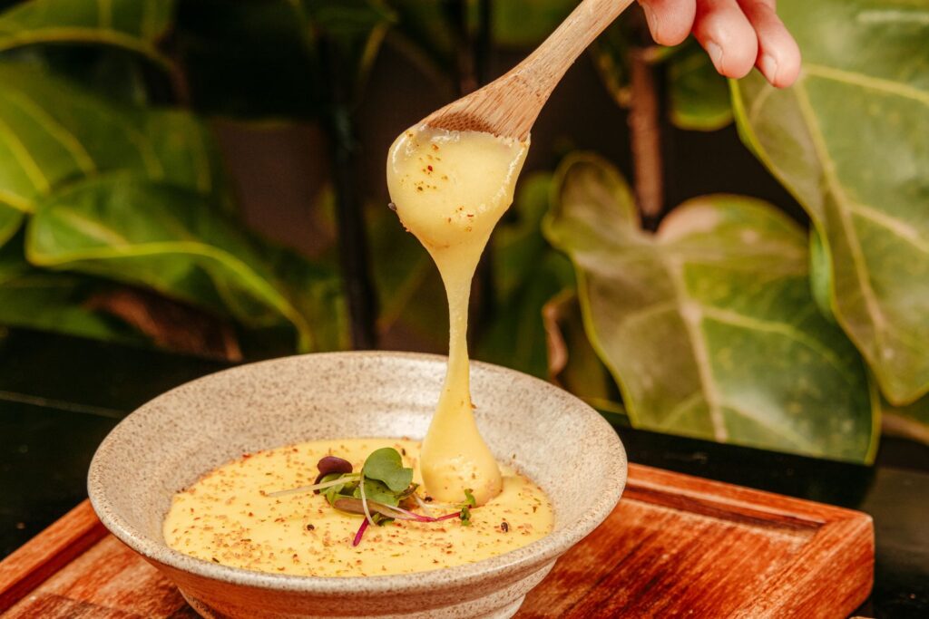 Spoon lifting creamy, cheesy yellow porridge from a rustic bowl, much like a serving of cheese grits, set against green leave