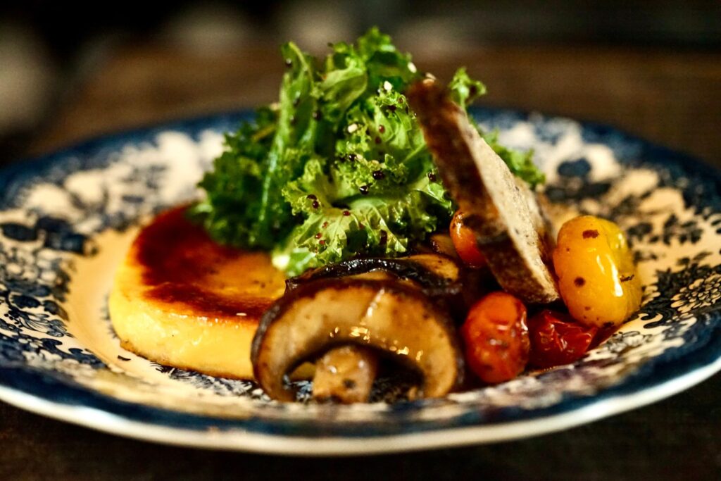 Seared slice of polenta served with roasted mushroom, cherry tomatoes and fresh greens on a blue floral plate