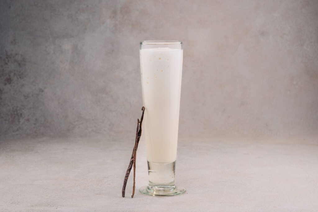 Tall glass of creamy white vanilla drink with a whole vanilla bean leaning against it, illustrating vanilla as a flavoring.